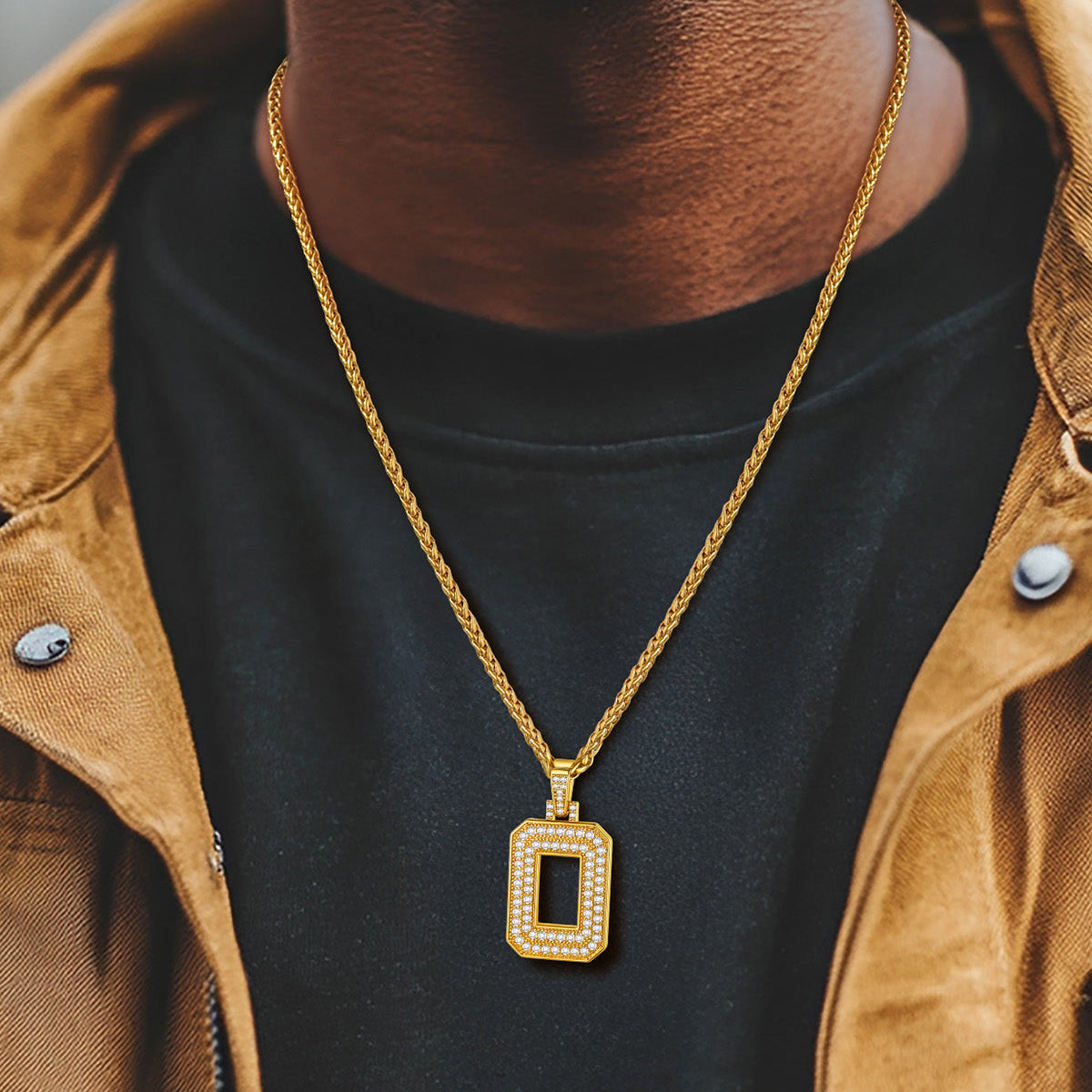 Person wearing a gold necklace with a rectangular pendant, dressed in a brown jacket and black shirt.