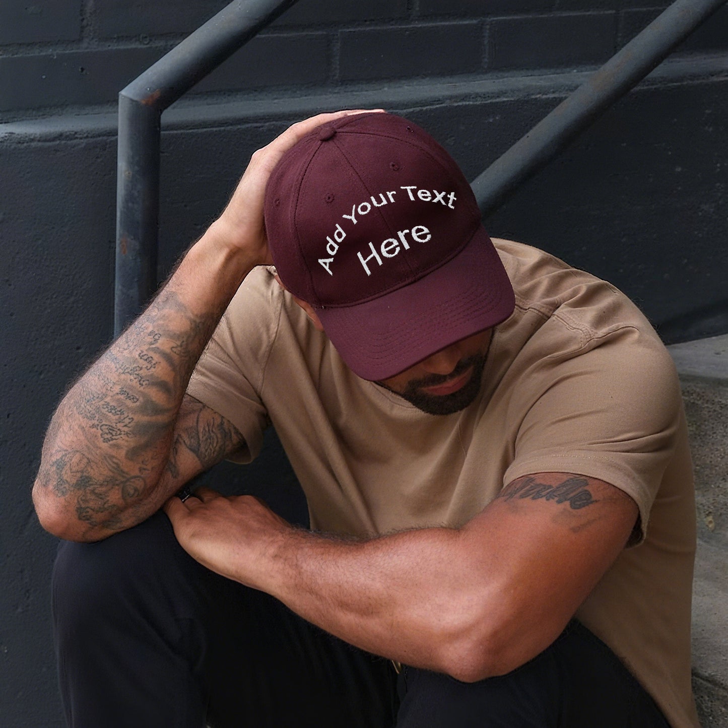 Man wearing a maroon cap with 'Add Your Text Here' text, sitting against a dark wall.