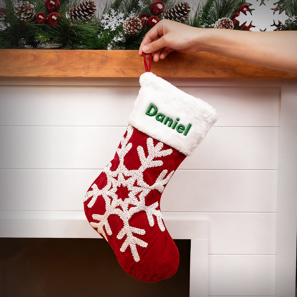 Red Christmas stocking with white snowflake pattern and green name 'Daniel' being hung on a fireplace mantle.