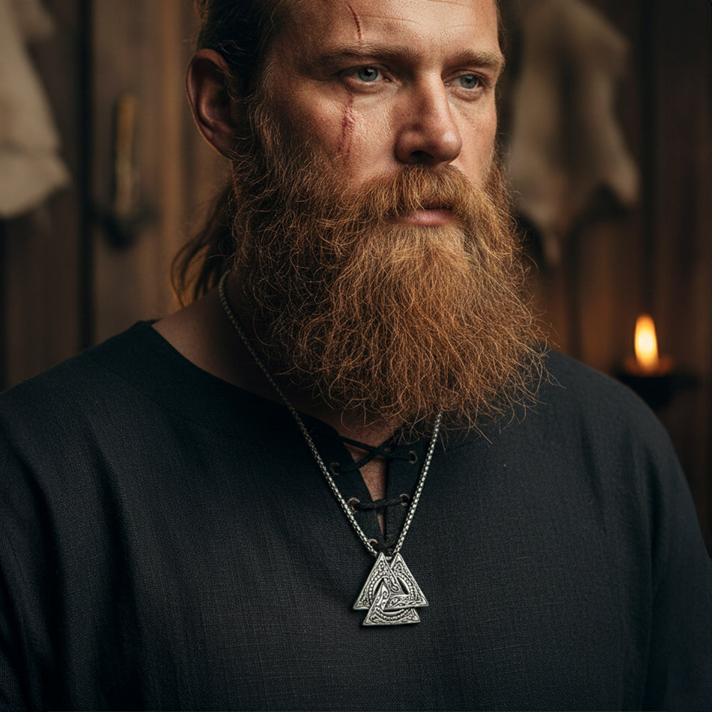 Man with a long beard wearing a dark shirt and a necklace with a triangular pendant, in a dimly lit room.