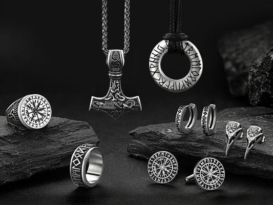 Collection of silver Viking jewelry displayed on dark stone surfaces, featuring Thor's hammer pendant, runic rings, circular compass pendants, earrings, and cufflinks with Norse symbols and inscriptions.