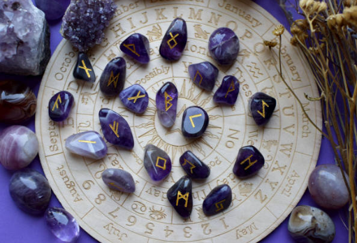 Purple amethyst rune stones with gold engravings arranged on a wooden circular zodiac board, surrounded by crystals and dried flowers on a purple background.