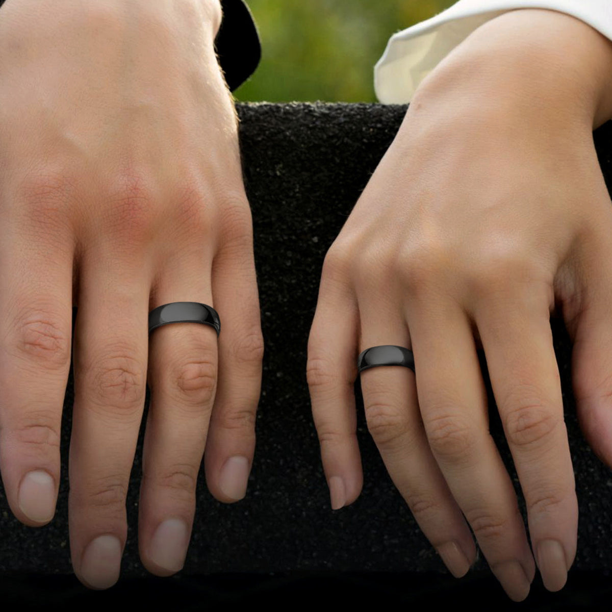 Couple resting on a dark textured surface, each wearing a simple matte black band ring. 
