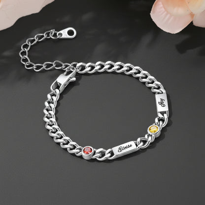 A silver Cuban chain bracelet with three engraved name plates, each set with a small round gemstone in red, yellow, and pink, displayed on a warm beige background.