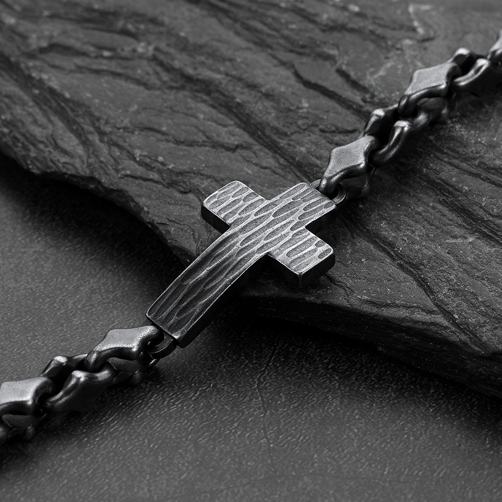 A dark metallic cross bracelet with a textured finish, attached to a matching chain, displayed on a dark slate surface.