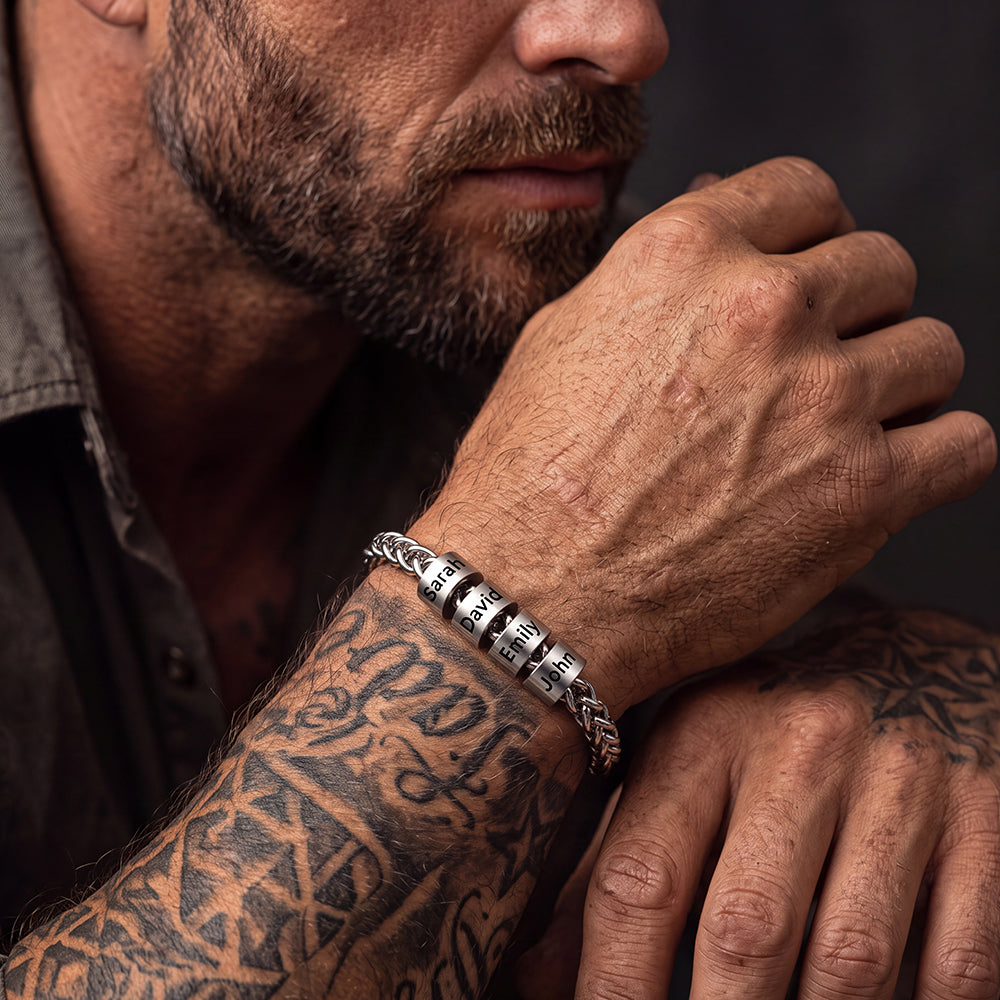 A close-up lifestyle shot of a man with tattoos wearing a silver-toned custom names bracelet on his wrist.