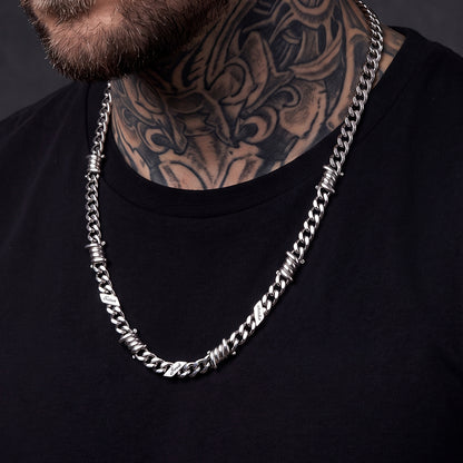 A close-up of a man wearing a thick silver-toned chain necklace with textured, twisted barbed wire details.