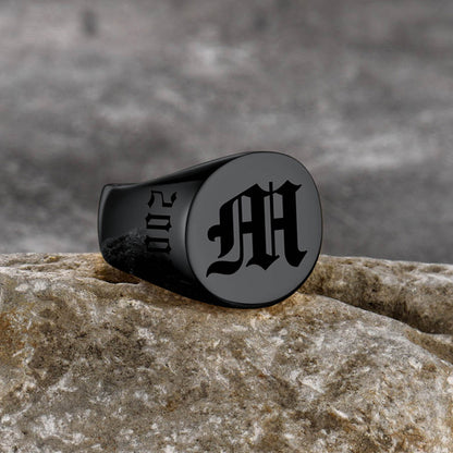 Black ring with engraved letter 'M' on a stone surface