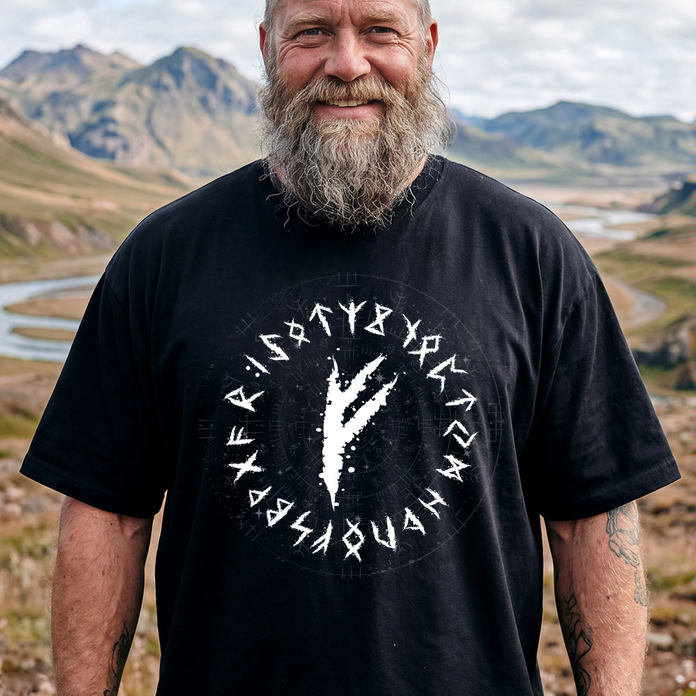 Man wearing a black t-shirt with a white viking rune design and text, standing in a scenic outdoor setting with mountains and a river.