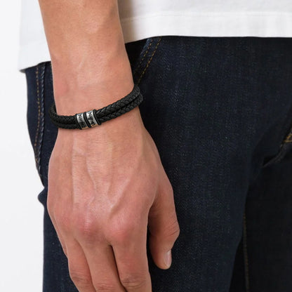 Hand wearing a black braided bracelet with silver accents, against a blurred background.