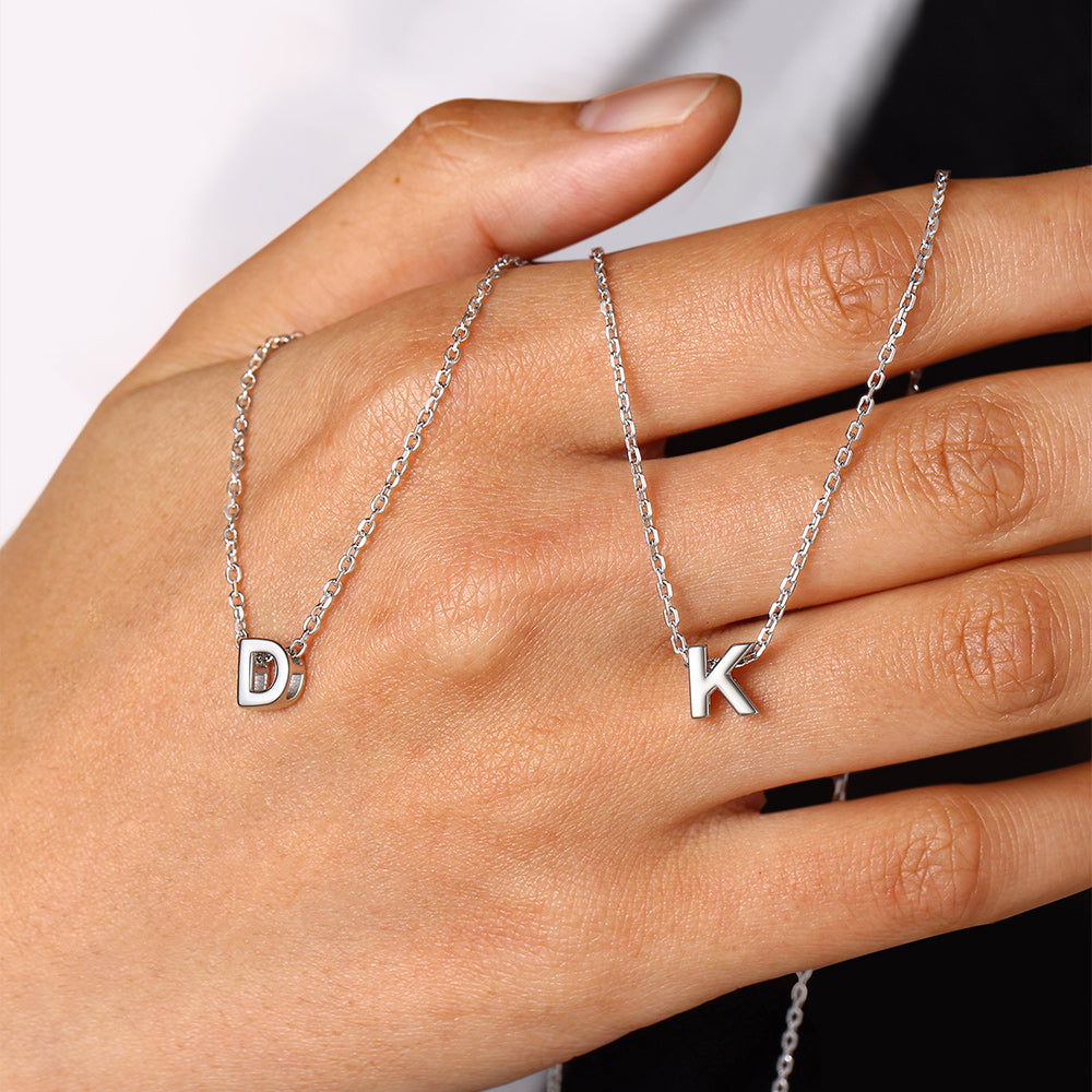 Hand wearing two silver necklaces with 'D' and 'K' charms on a neutral background