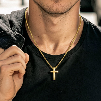A close-up lifestyle shot of a man wearing a gold cross pendant necklace on a black t-shirt, with a blurred outdoor background.