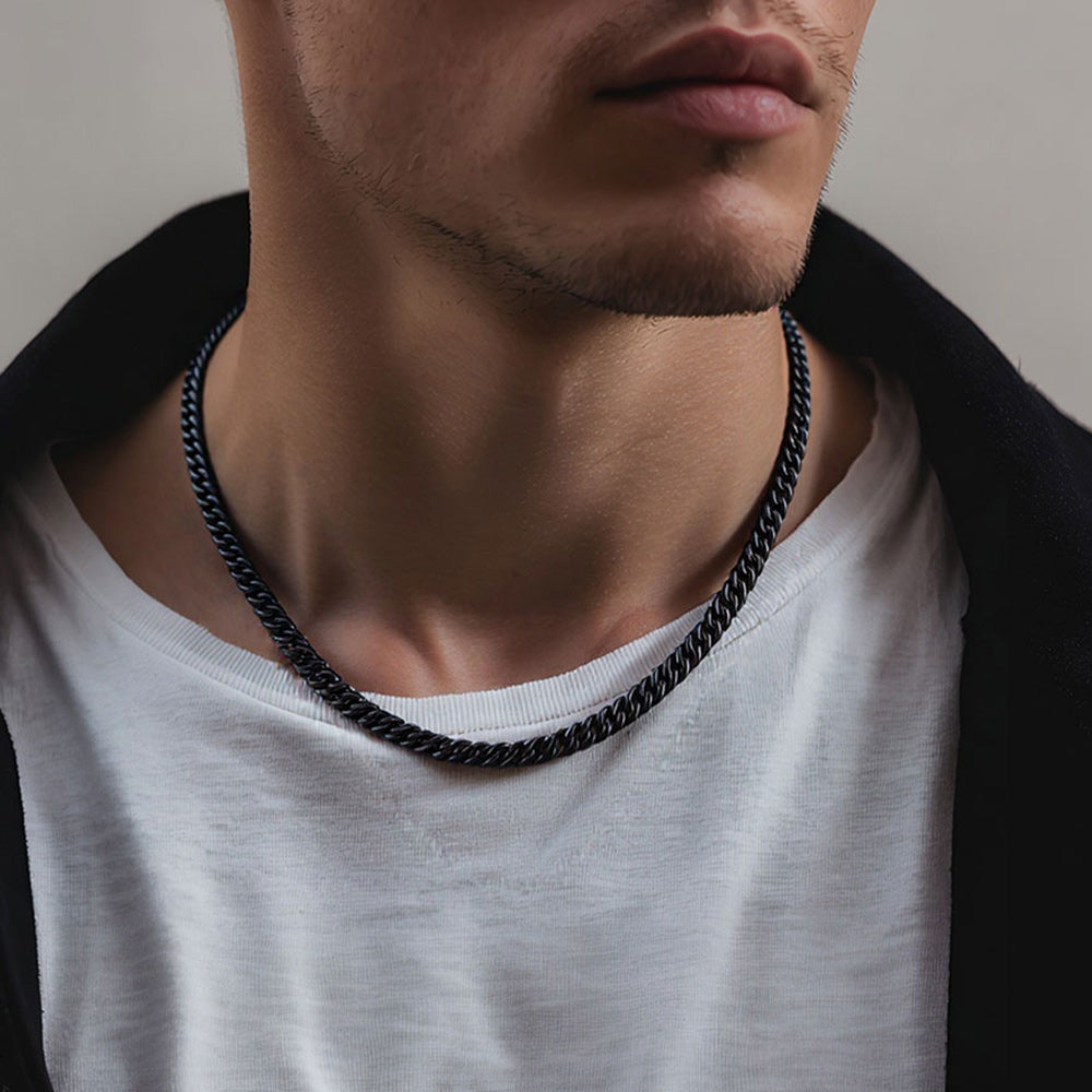 A close-up of a man's neck and lower face wearing a thick black twisted cuban chain necklace over a white t-shirt and dark jacket.
