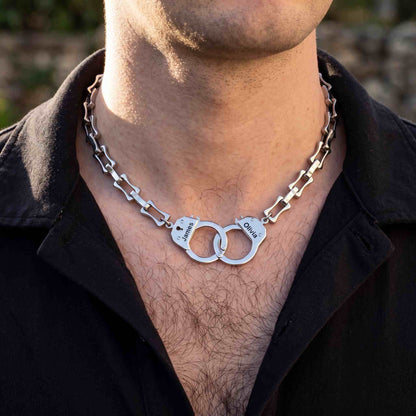 A silver metal chain necklace shaped like handcuffs, worn by a man in a black shirt. 