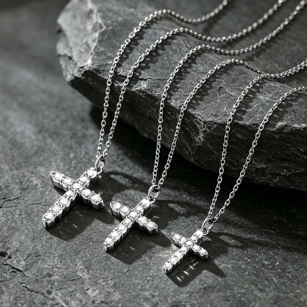 Three silver cross pendant necklaces with clear stones, arranged on a dark textured stone surface.