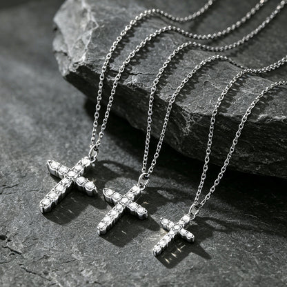 Three silver cross pendant necklaces with clear stones, arranged on a dark textured stone surface.