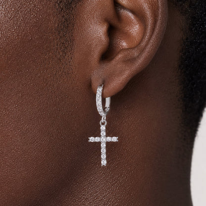 A close-up of a person's ear wearing a silver huggie hoop earring with a dangling cross charm, both set with small clear stones.
