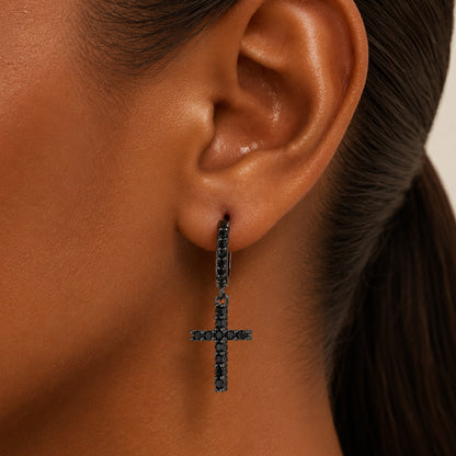 A close-up of a person's ear wearing a small cross drop earring with dark stones.