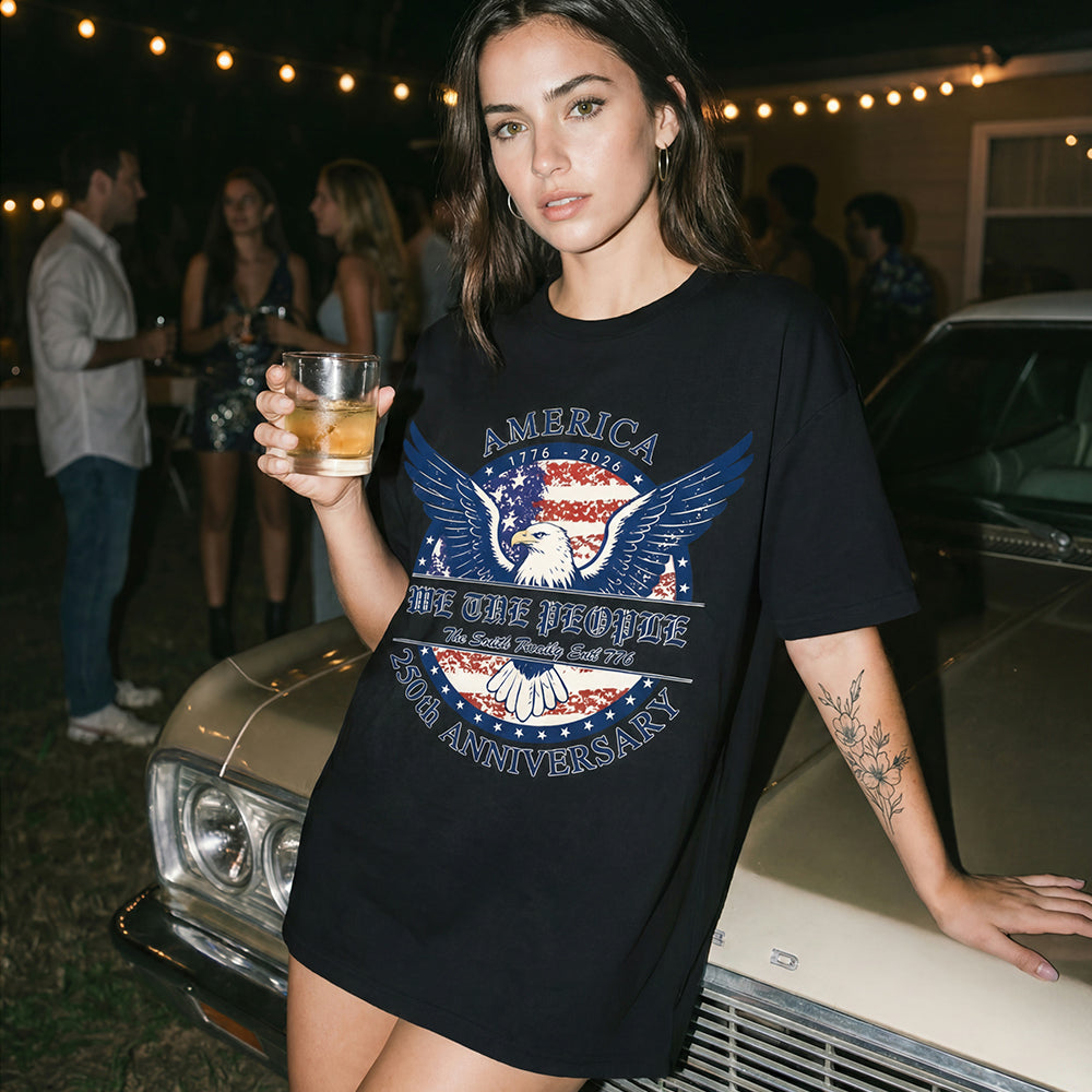 Woman in a black t-shirt with an eagle graphic and text, holding a drink, standing next to a vintage car at a party.