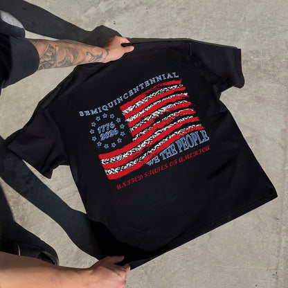Black t-shirt with American flag design and text held by a person on a concrete surface