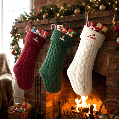 Three personalized cable-knit Christmas stockings hang from a brick fireplace with a fire burning below. 