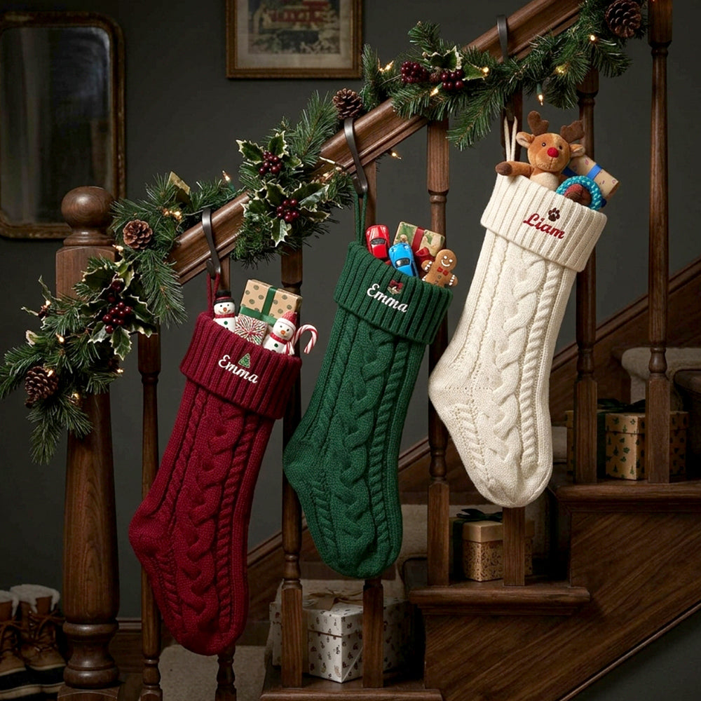 Three personalized Christmas cable-knit stockings hang from a wooden staircase railing decorated with a green garland. 