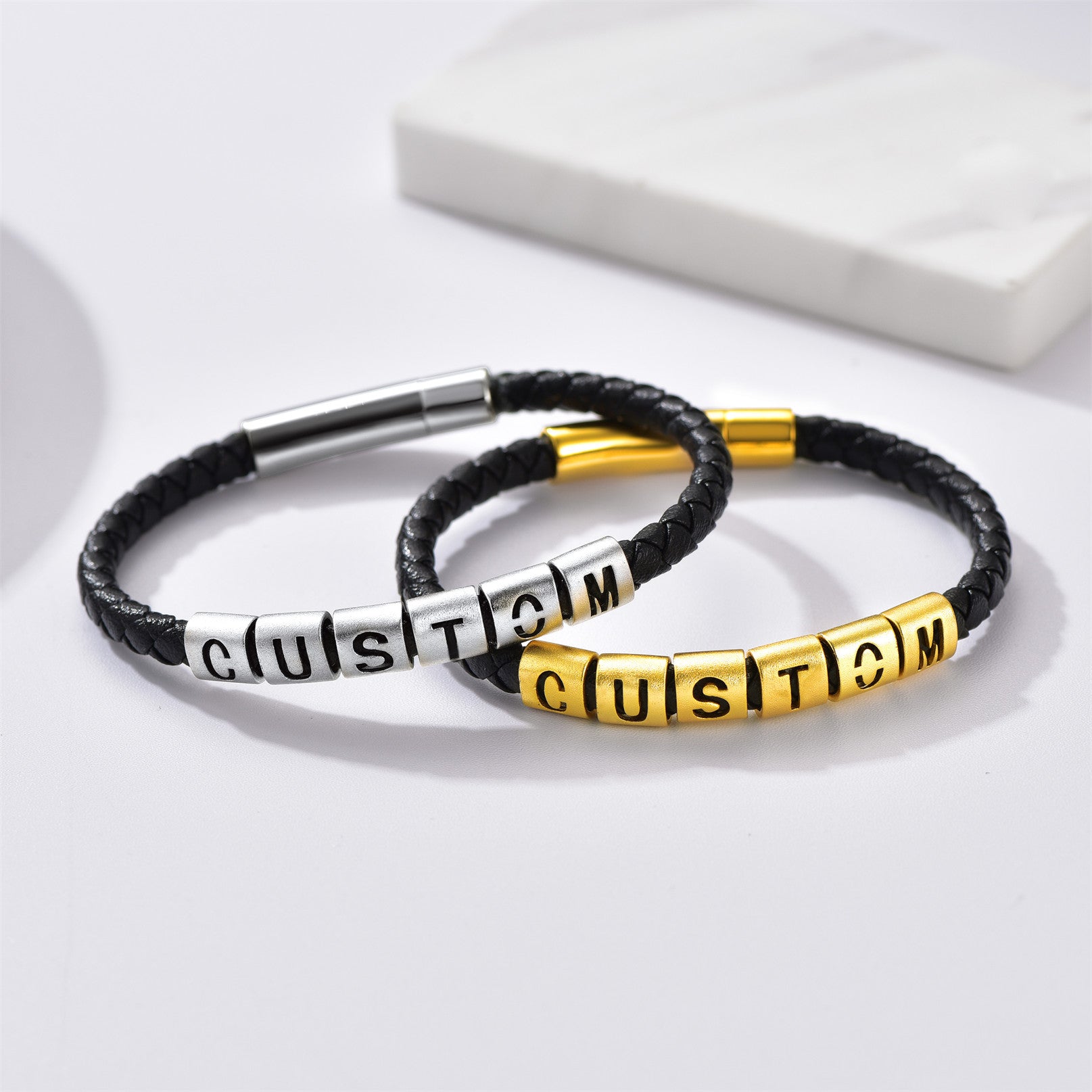 Two black leather bracelets with metal beads spelling 'CUSTOM' on a white background