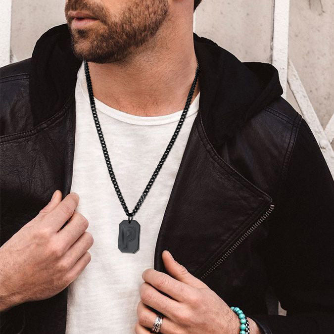 A man wearing a black metal chain necklace with a rectangular pendant featuring a stylized letter 'P', styled over a white t-shirt and black leather jacket.