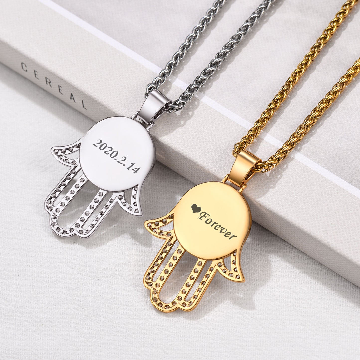 Two Hamsa hand pendants, one silver and one gold, on a white surface.