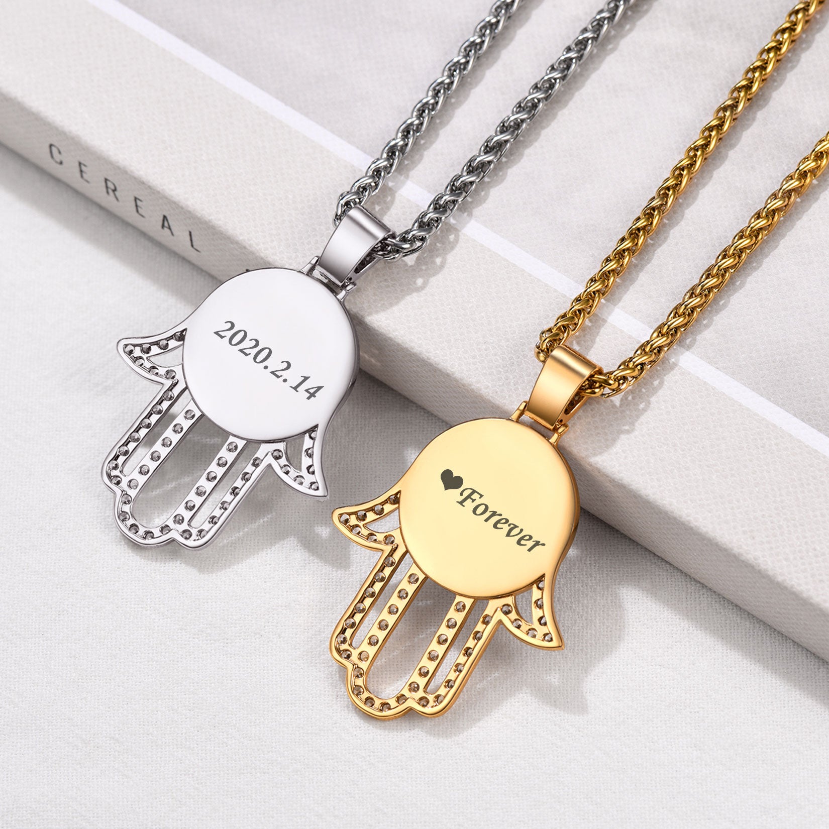 Two Hamsa hand pendants, one silver and one gold, on a white surface.