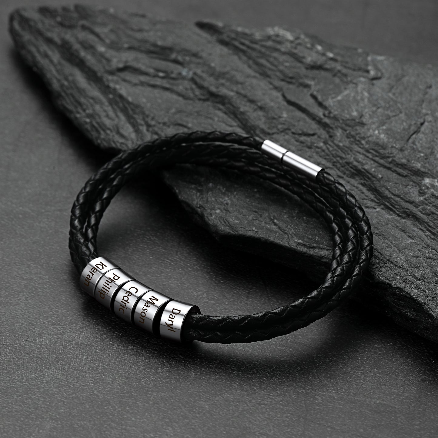 Black braided leather bracelet with metal tags on a dark stone surface