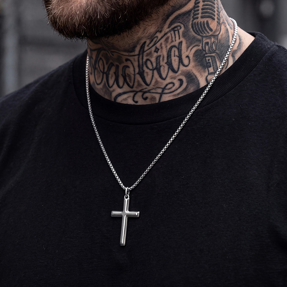 A silver cross pendant necklace on a silver chain, worn by a man with a beard and neck tattoos wearing a black t-shirt.