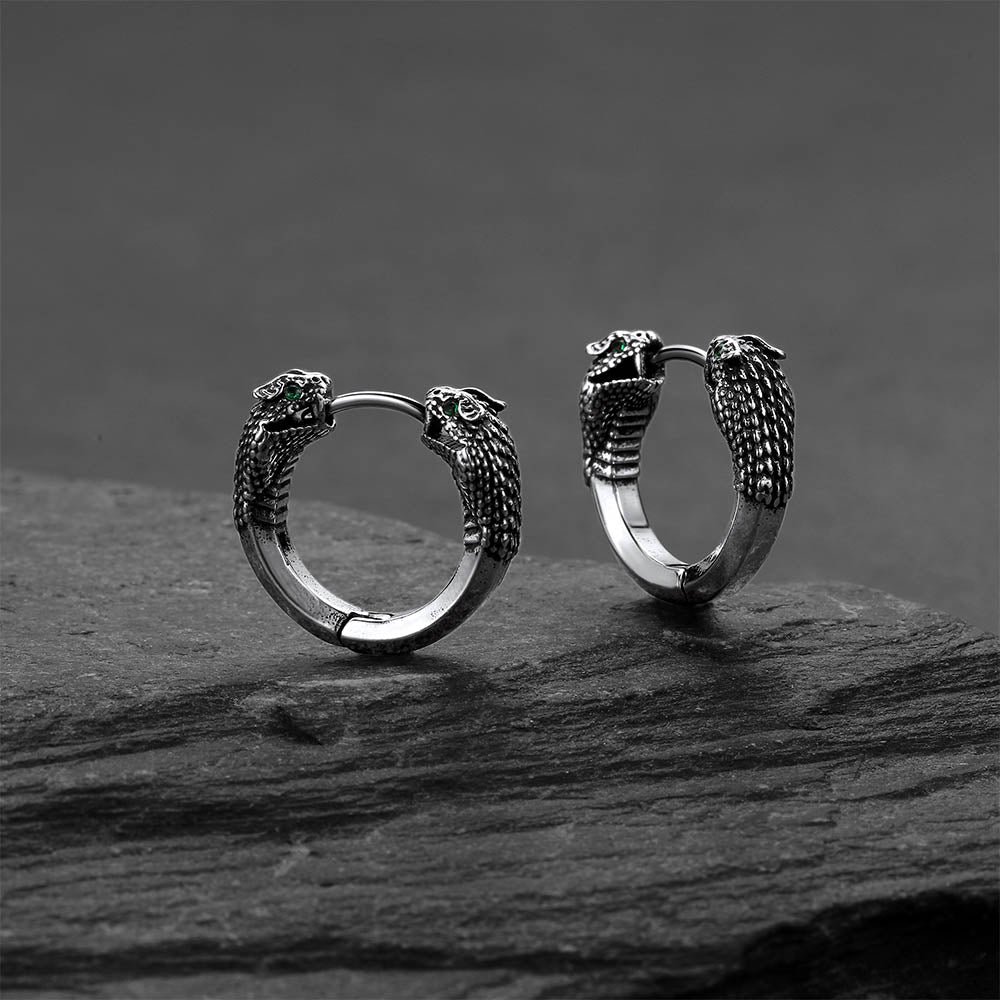 Pair of silver hoop earrings with snake head designs on a dark stone surface.