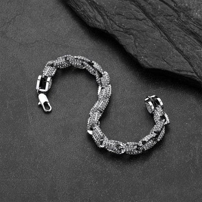 A silver-toned chain link bracelet with pavé-set clear stones, arranged in a curved S-shape on a dark textured slate surface with a larger dark stone in the upper right corner.
