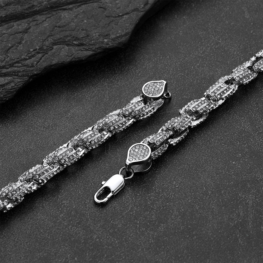 A silver-toned Cuban link chain bracelet with pavé-set clear stones, shown in two pieces on a dark textured surface. One piece displays the full chain with a heart-shaped pavé clasp and lobster clasp, while the other shows a close-up of the clasp detail.