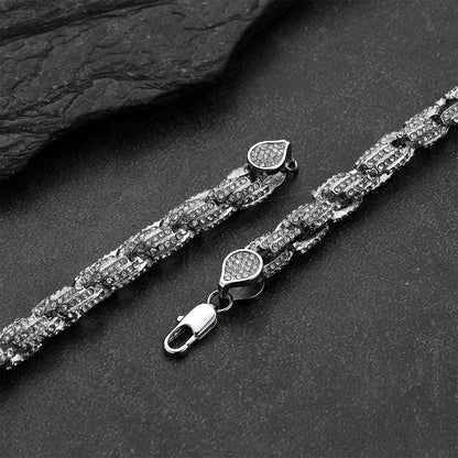 A silver-toned Cuban link chain bracelet with pavé-set clear stones, shown in two pieces on a dark textured surface. One piece displays the full chain with a heart-shaped pavé clasp and lobster clasp, while the other shows a close-up of the clasp detail.