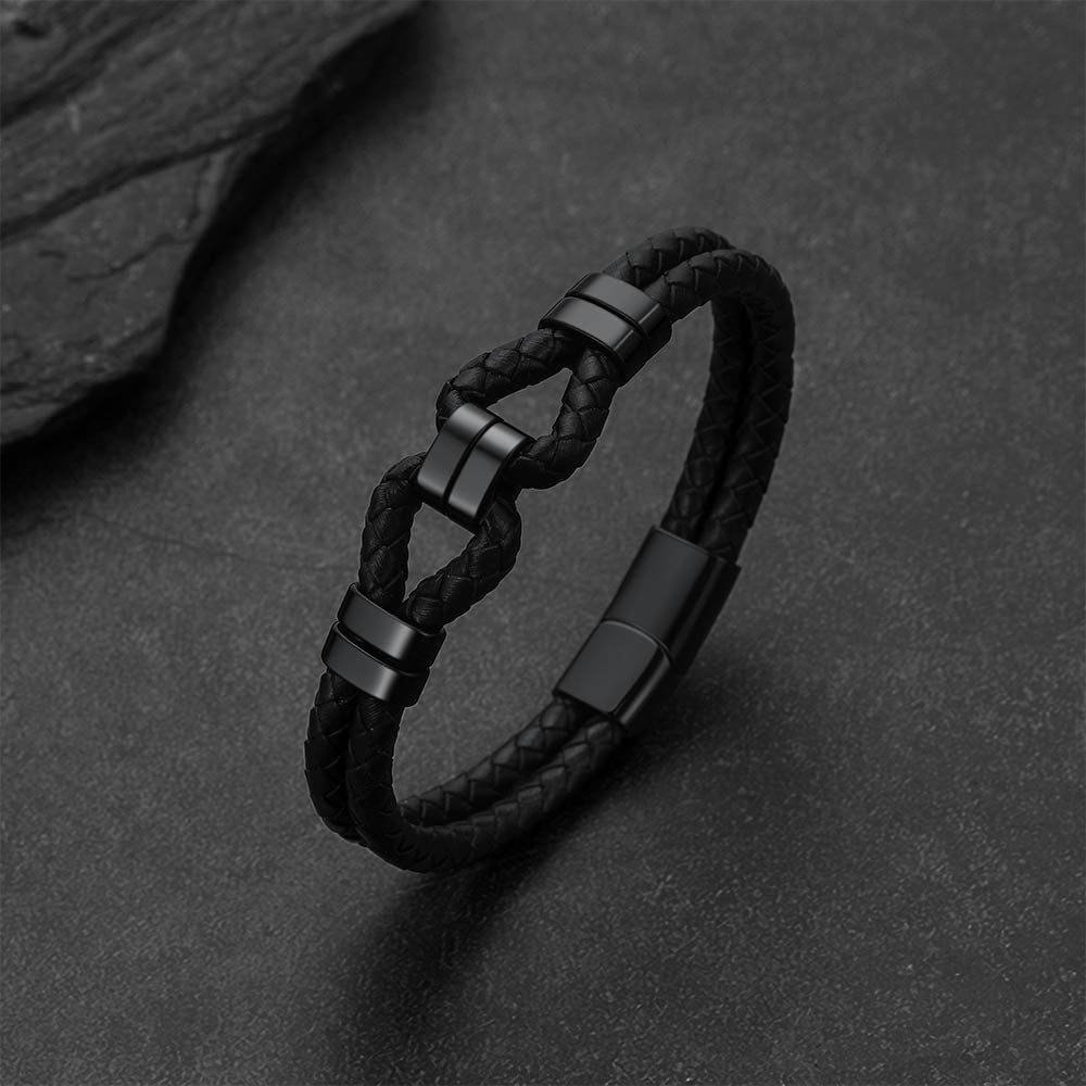 A black braided leather bracelet with matte black metal connector pieces, displayed on a dark textured surface.