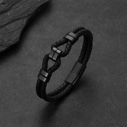 A black braided leather bracelet with matte black metal connector pieces, displayed on a dark textured surface.