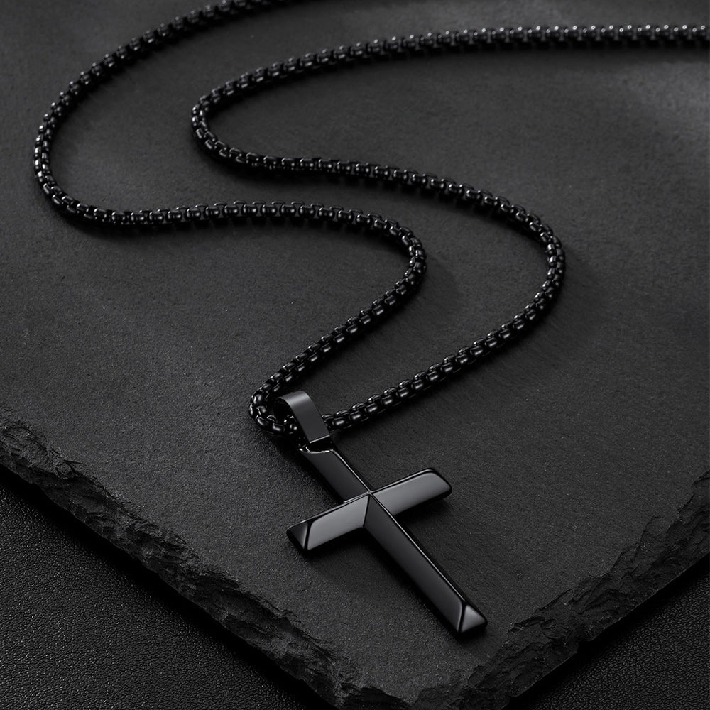 A black beveled edge cross pendant necklace displayed on a dark textured slate surface.