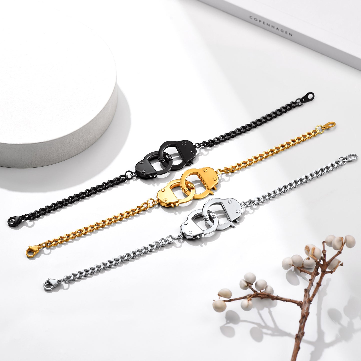 Three chain bracelets in black, gold, and silver on a white surface with a branch and cylindrical object.