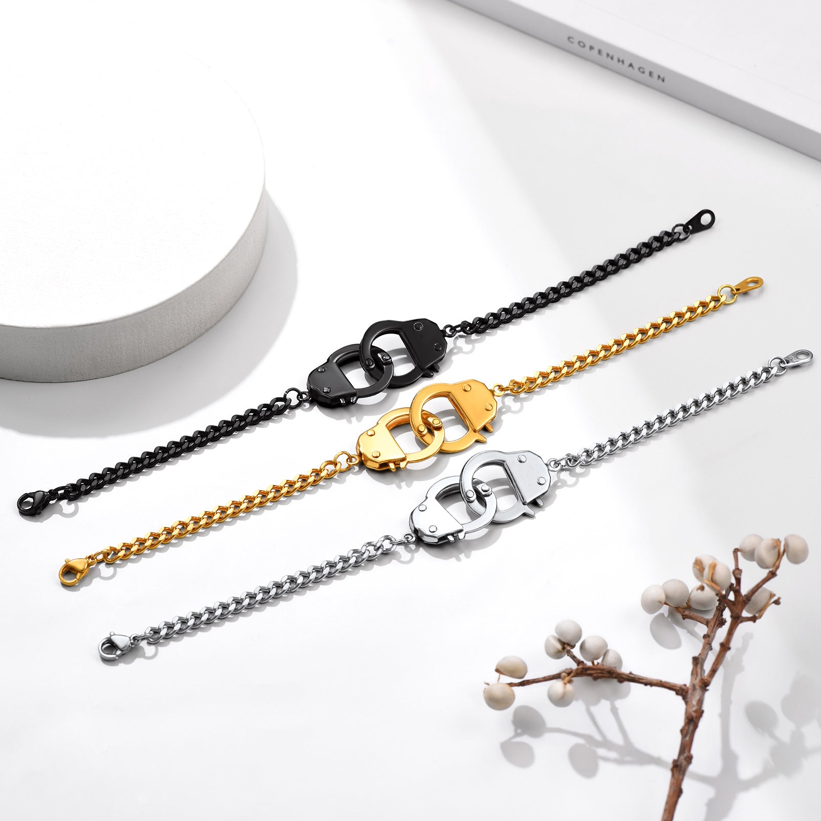 Three chain bracelets in black, gold, and silver on a white surface with a branch and cylindrical object.