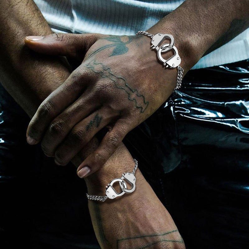 Two hands with tattoos wearing silver handcuff bracelets against a dark background.