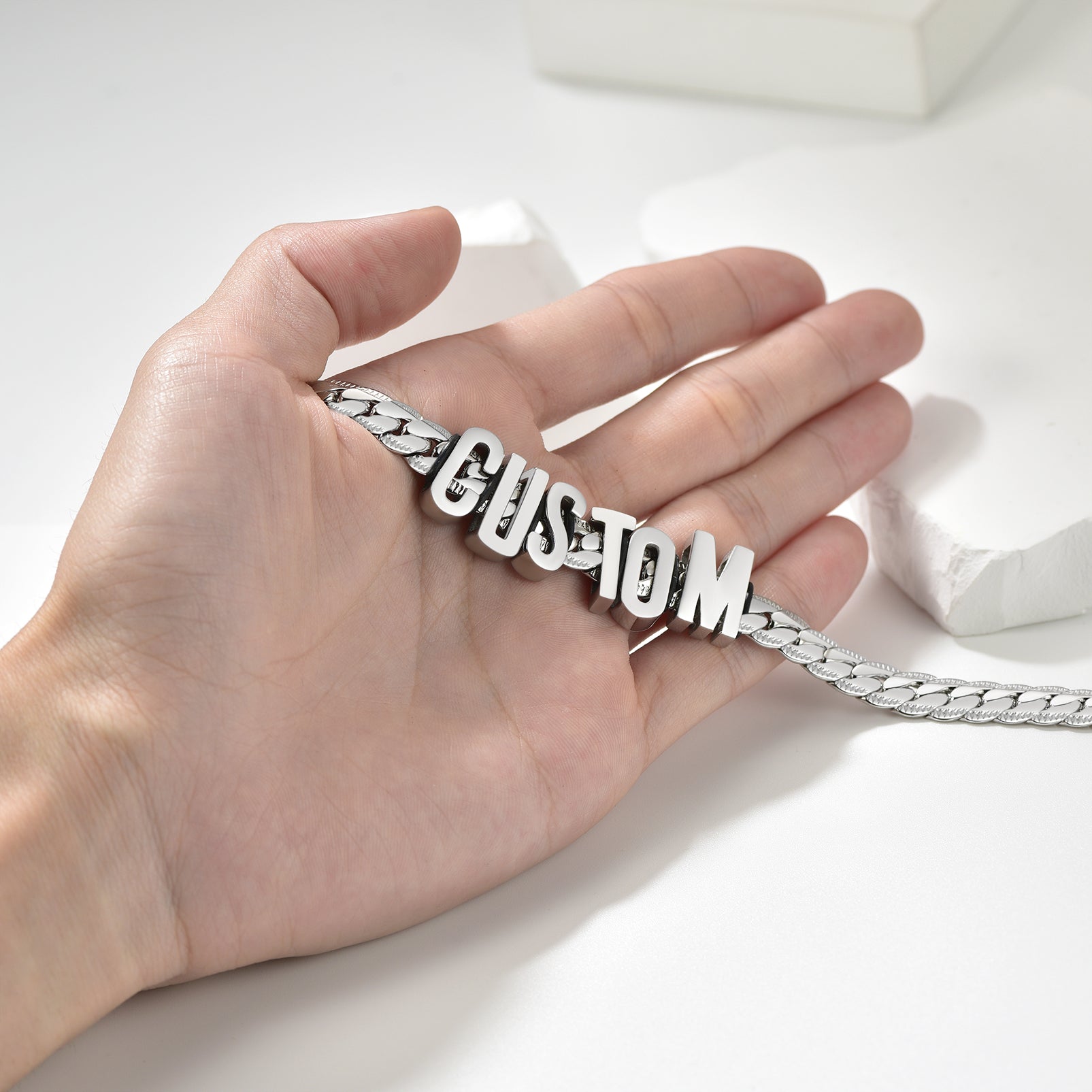 Hand holding a silver chain with 'CUSTOM' letters on a white background