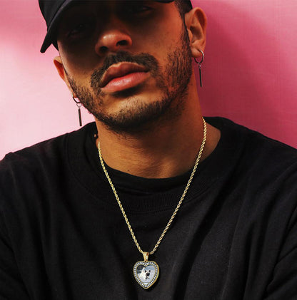 Man wearing a black shirt and gold necklace with a heart-shaped pendant against a pink background
