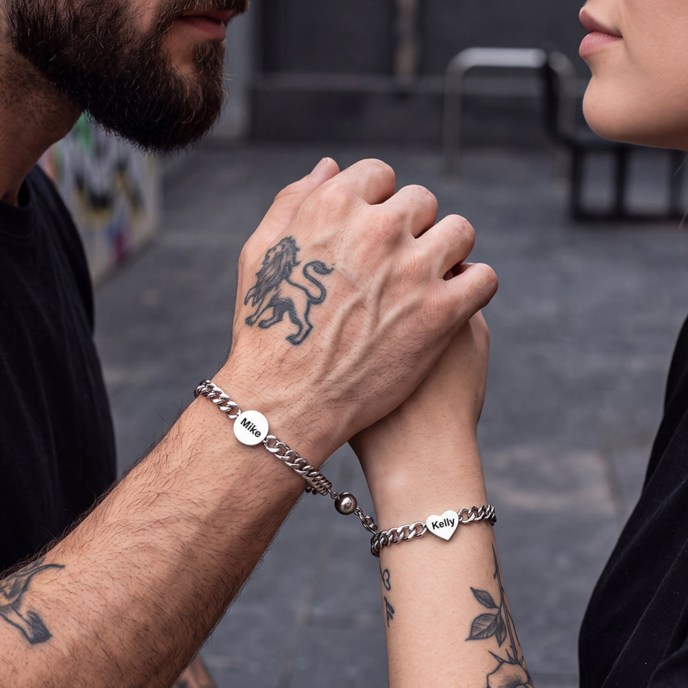Two people each wearing a silver chain bracelet with a small heart-shaped charm engraved with a name.