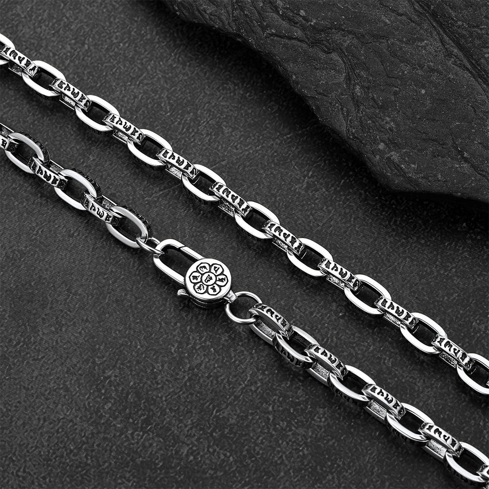 A close-up of a silver-toned metal chain necklace with elongated oval links and a lobster clasp, laid diagonally on a dark textured surface with a black stone slab in the background.