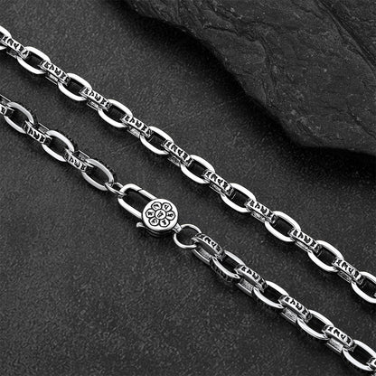 A close-up of a silver-toned metal chain necklace with elongated oval links and a lobster clasp, laid diagonally on a dark textured surface with a black stone slab in the background.