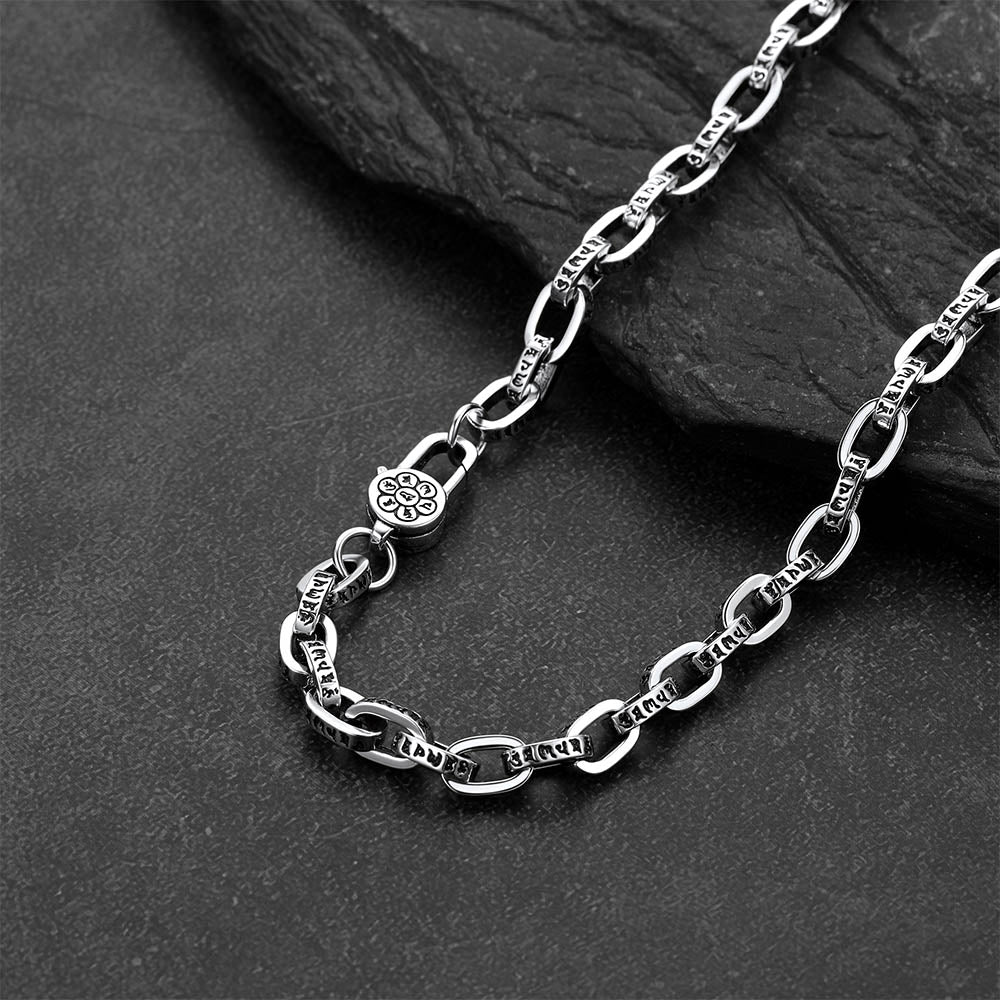 A close-up of a silver-toned metal chain necklace with textured oval links and a decorative clasp, arranged on a dark stone surface.