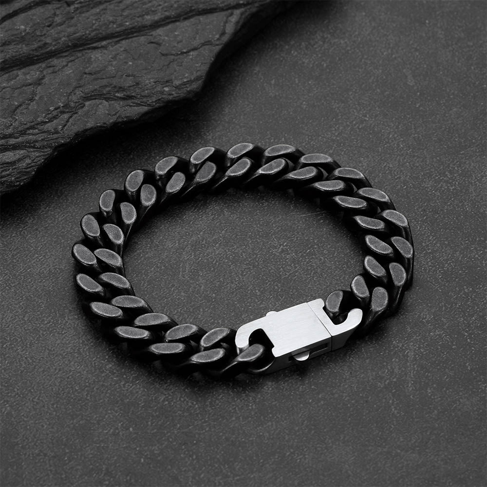 A black metal curb link bracelet, displayed on a dark textured slate surface.