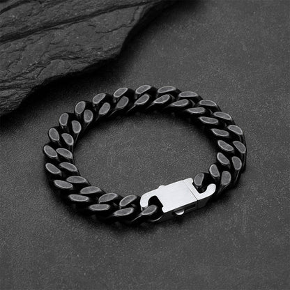 A black metal curb link bracelet, displayed on a dark textured slate surface.